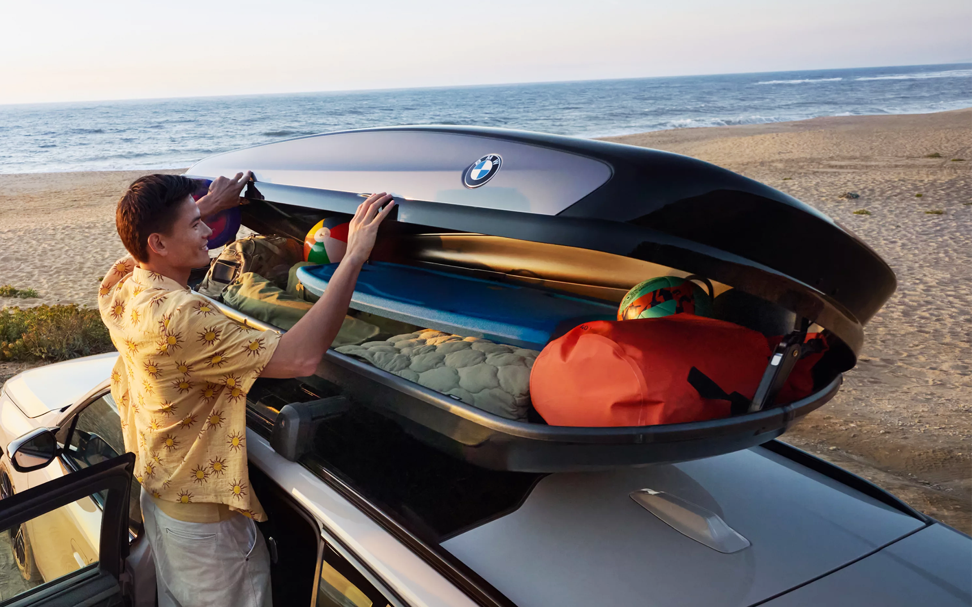 Un homme ouvre un coffre de toit BMW rempli d’accessoires de plage, avec la mer en arrière-plan.